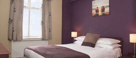Una habitación de hotel con una cama grande, dos lámparas de noche, un ventanal con cortinas y un cuadro en la pared.