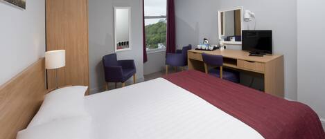 A hotel room with a large bed, a desk with a computer, a chair, a lamp, and a window with curtains.