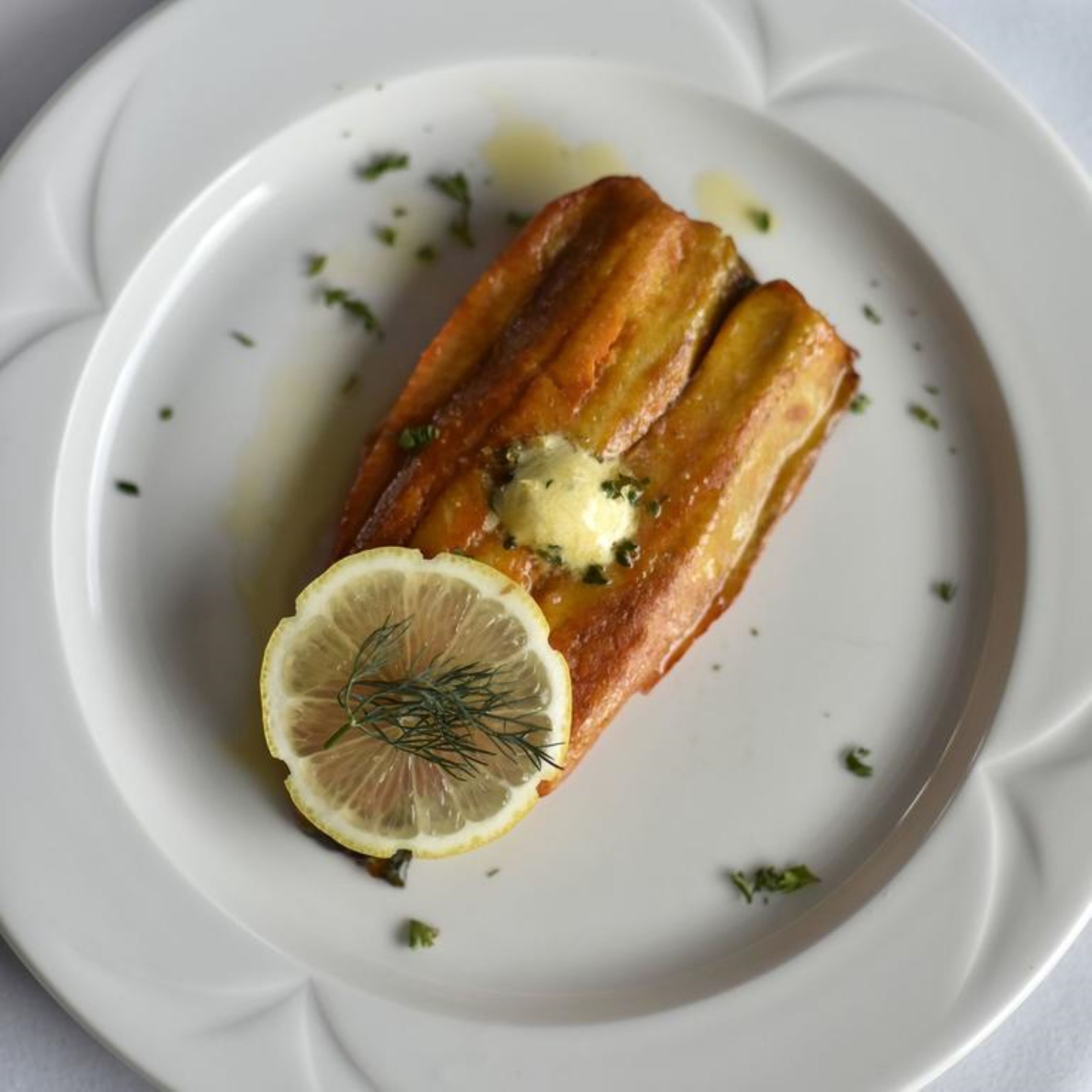 Un plato con un trozo de comida, posiblemente pescado, cubierto de mantequilla y adornado con una rodaja de limón y hierbas.