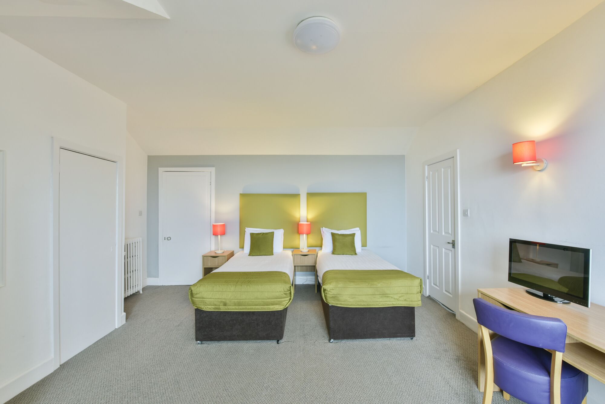 A hotel room with two beds, a desk, a chair, a television, and a wall-mounted lamp.