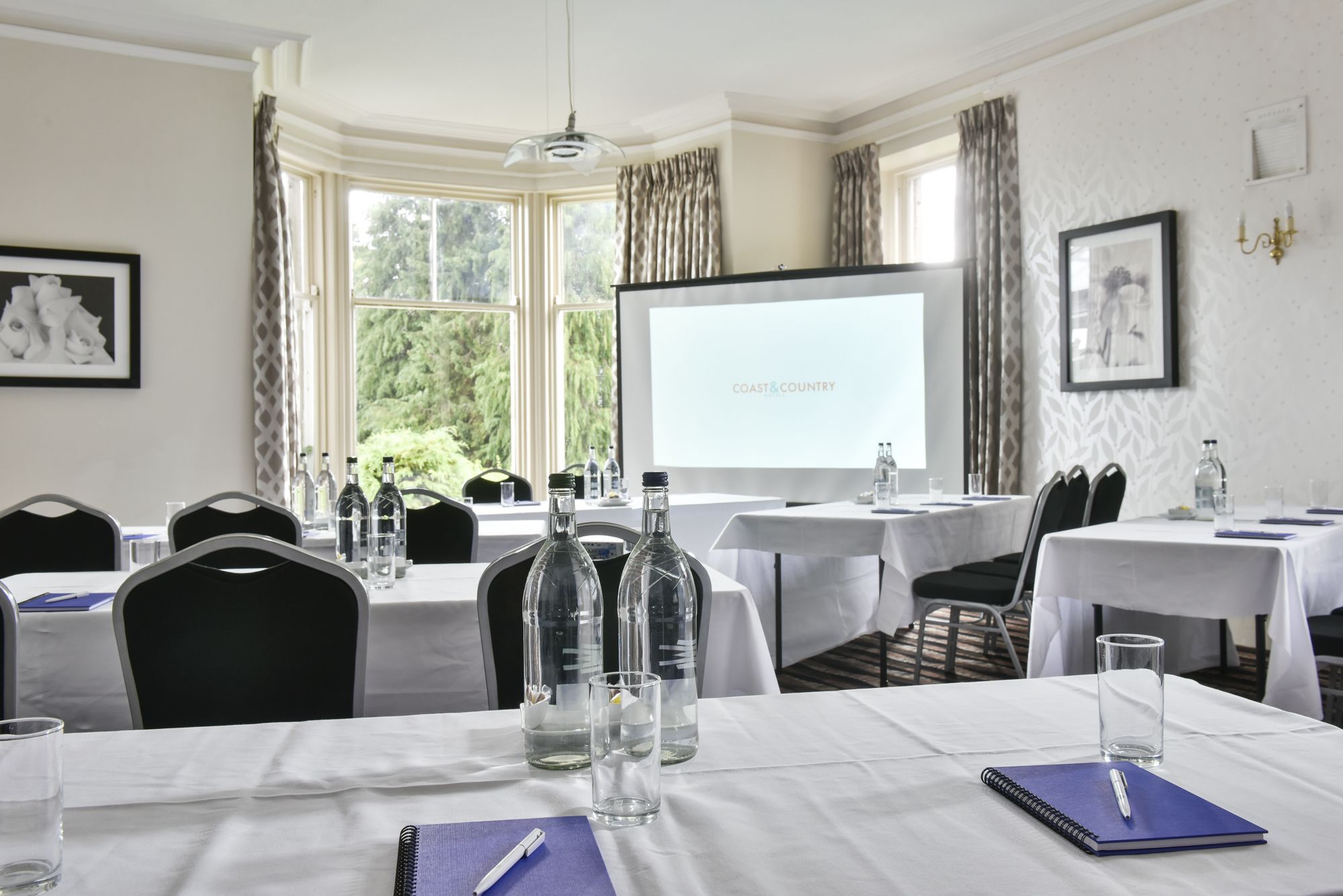 A conference room with tables set for a meeting, a projector screen displaying "COAST & COUNTRY", and a view of greenery outside.