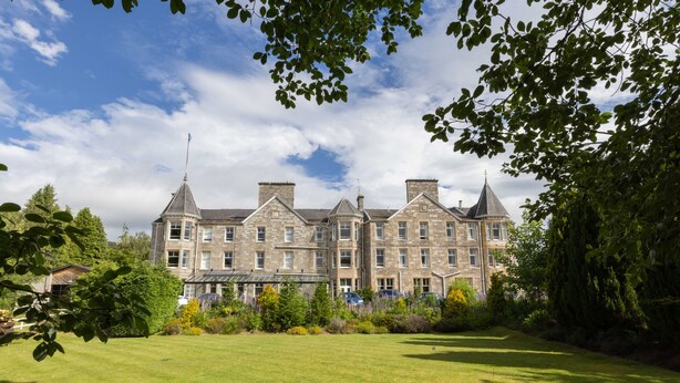 A large, historic building with multiple chimneys and a prominent tower, surrounded by a well-maintained lawn and trees. - The Pitlochry Hydro Hotel (Pitlochry)