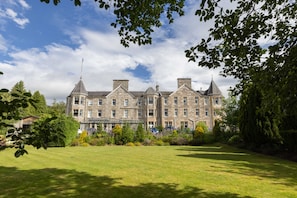A large, historic building with multiple chimneys and a prominent tower, surrounded by a well-maintained lawn and trees. - The Pitlochry Hydro Hotel (Pitlochry)