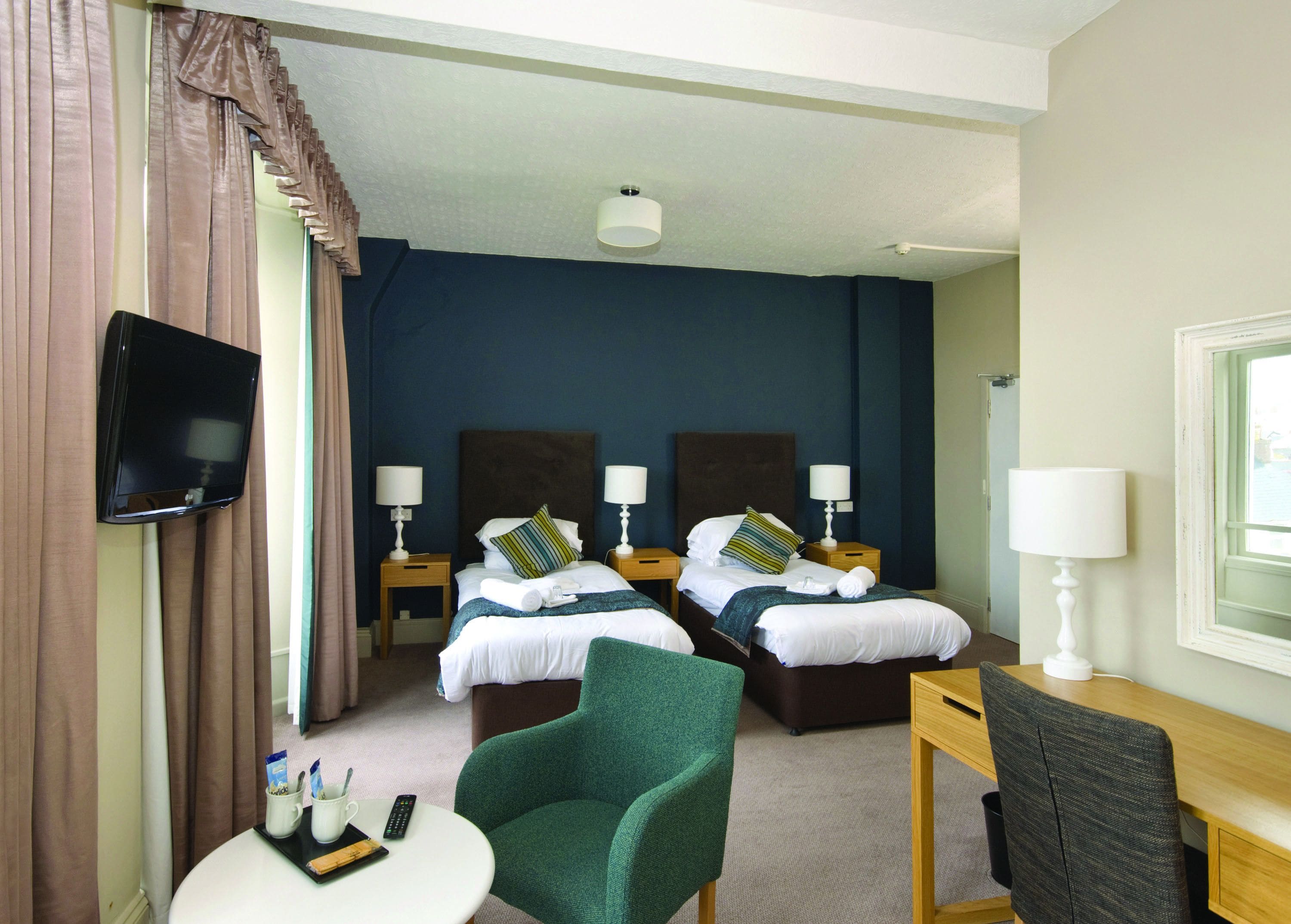 A hotel room with two beds, a TV mounted on the wall, a desk with a chair, and a window.