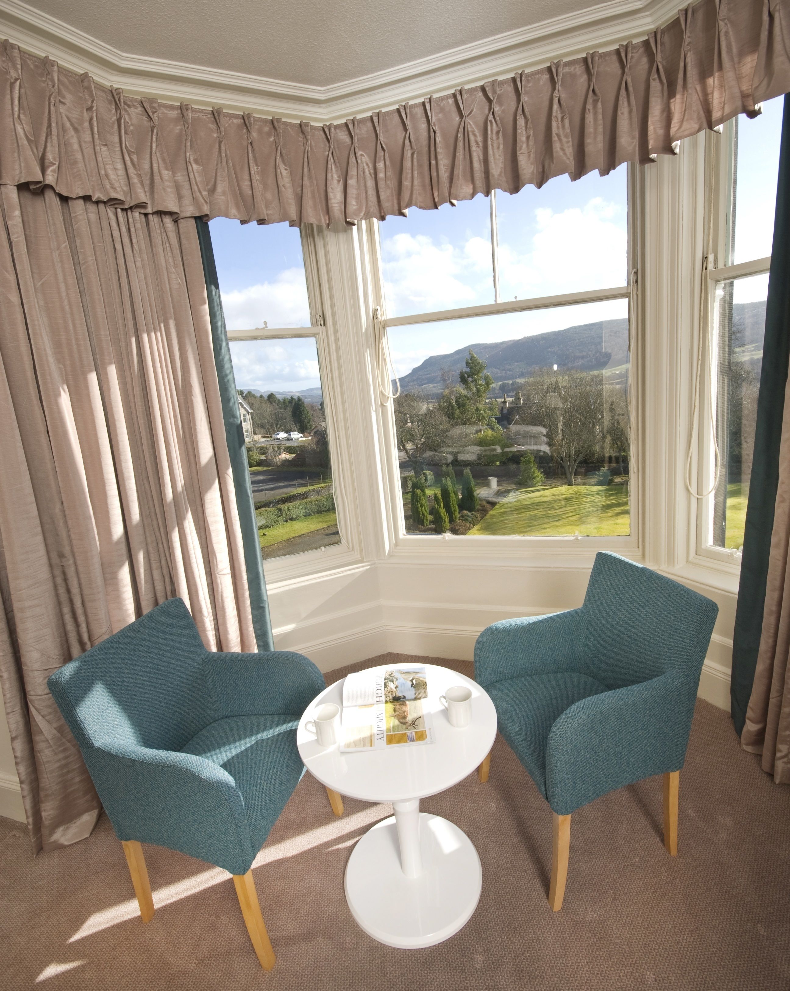 A room with two teal chairs, a white table, and a view of a landscape through large windows.