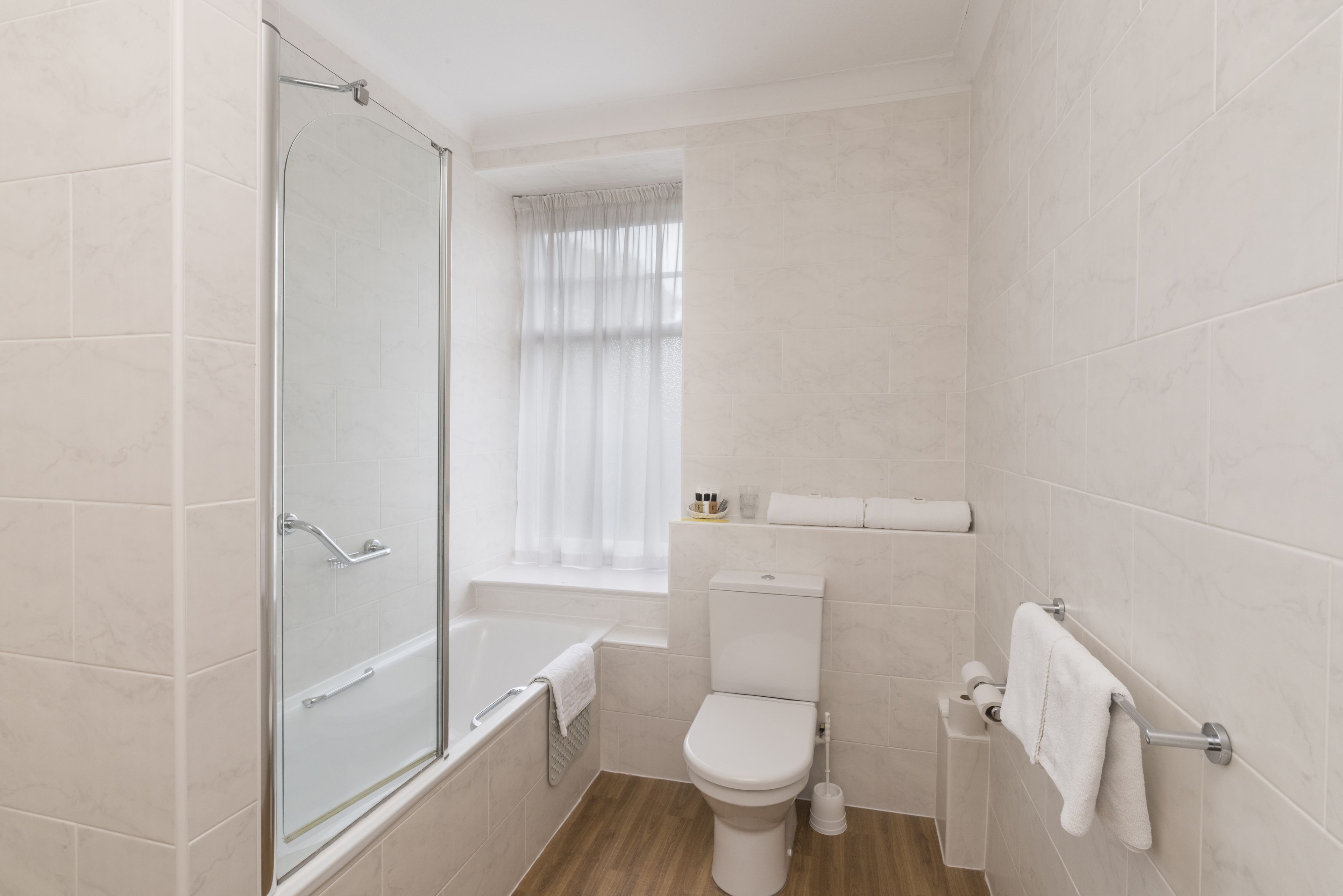 A bathroom with a glass shower enclosure, a toilet, a bathtub, and a window with curtains.