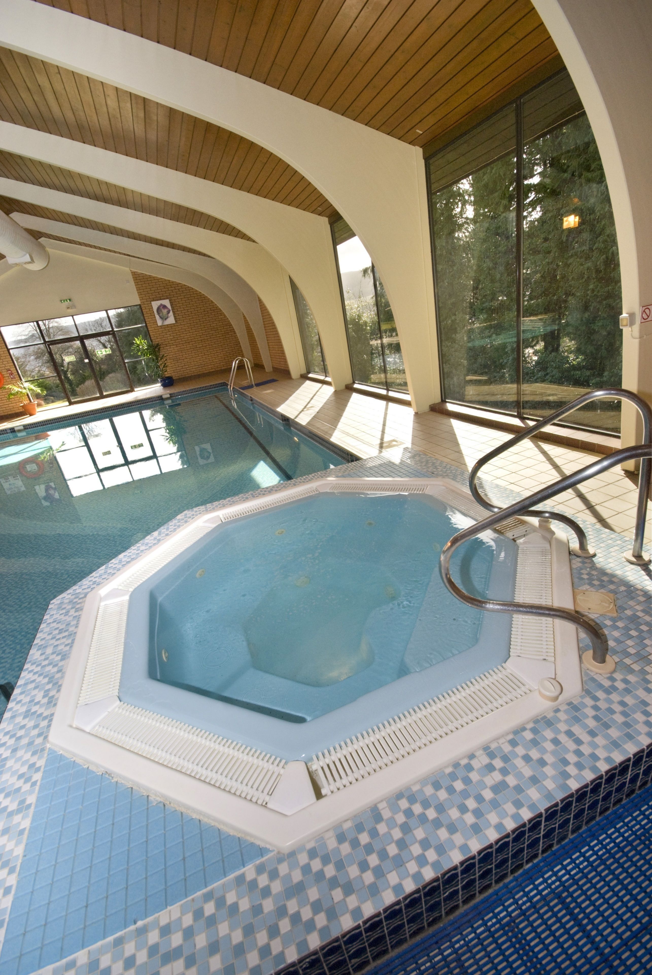 Una piscina interior con jacuzzi, suelo de baldosas y vistas a árboles a través de amplios ventanales.