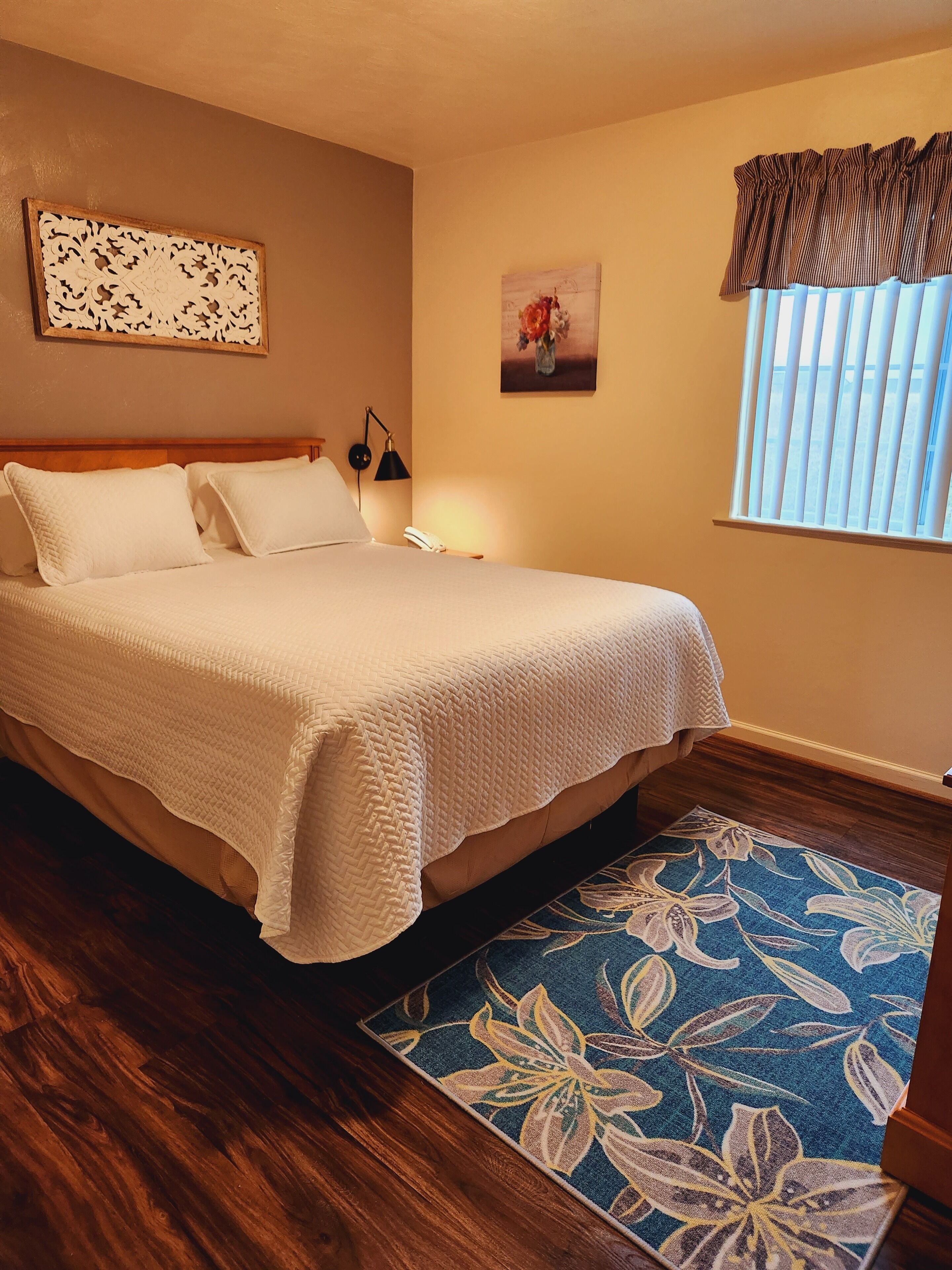 A neatly made bed with a white blanket, a wooden headboard, a wall-mounted lamp, a floral painting, and a window with blinds.