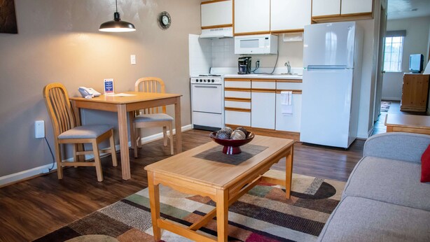 Een kleine keuken met witte kastjes, een magnetron, een fornuis en een koelkast. Er staat een eettafel met stoelen en een klein vloerkleed.