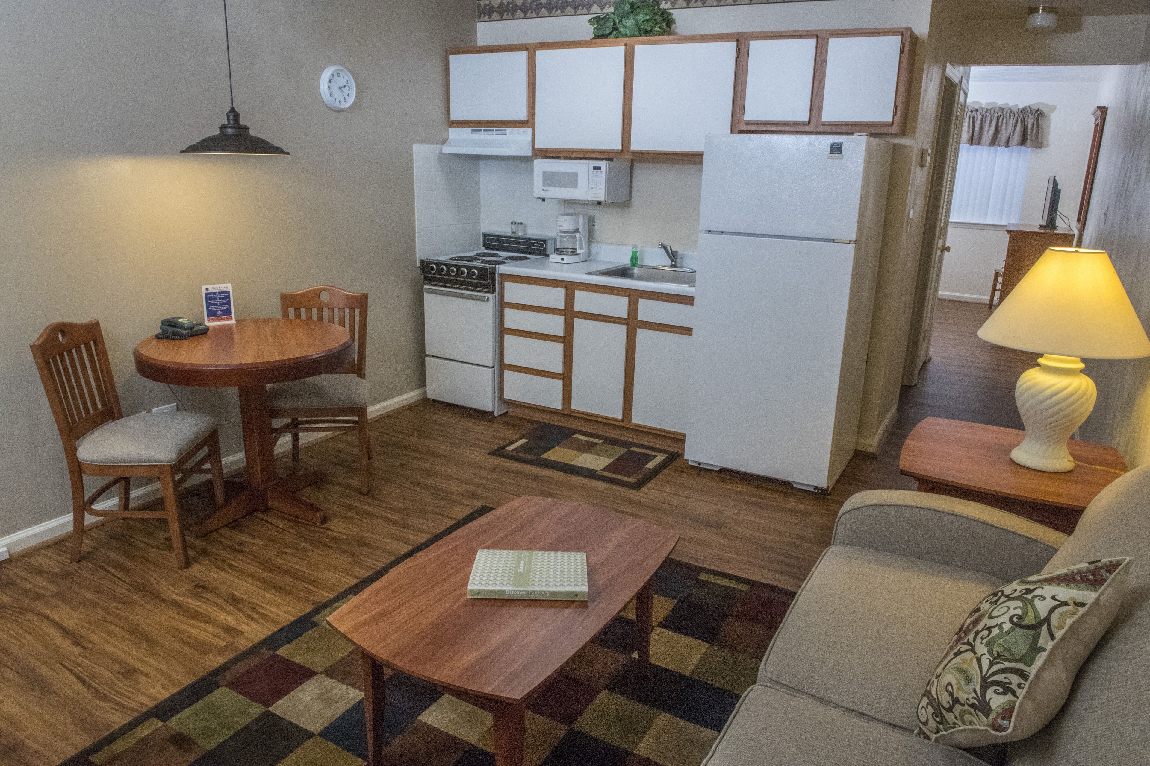 Een ruimtelijk ingerichte leefruimte met een kitchenette, een eethoek met tafel en stoelen, een bank met kussen, en een lamp op een bijzettafeltje.