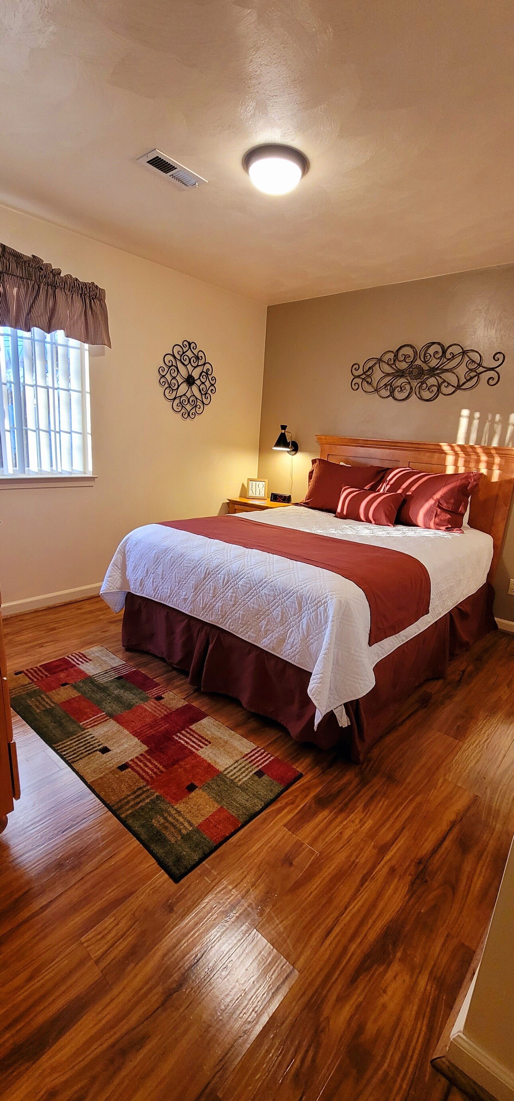 Un dormitorio con suelo de madera, una cama con colcha roja y blanca, una mesita de noche con lámpara y decoración mural.