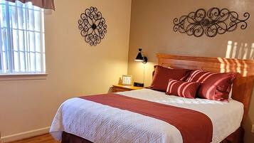 Un dormitorio con suelo de madera, una cama con colcha roja y blanca, una mesita de noche con lámpara y decoración mural.
