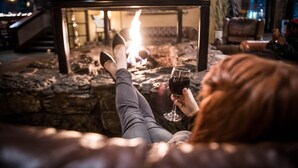 A person relaxing by a stone fireplace with a glass of red wine, in a cozy indoor setting with a stone wall and a staircase in the background.