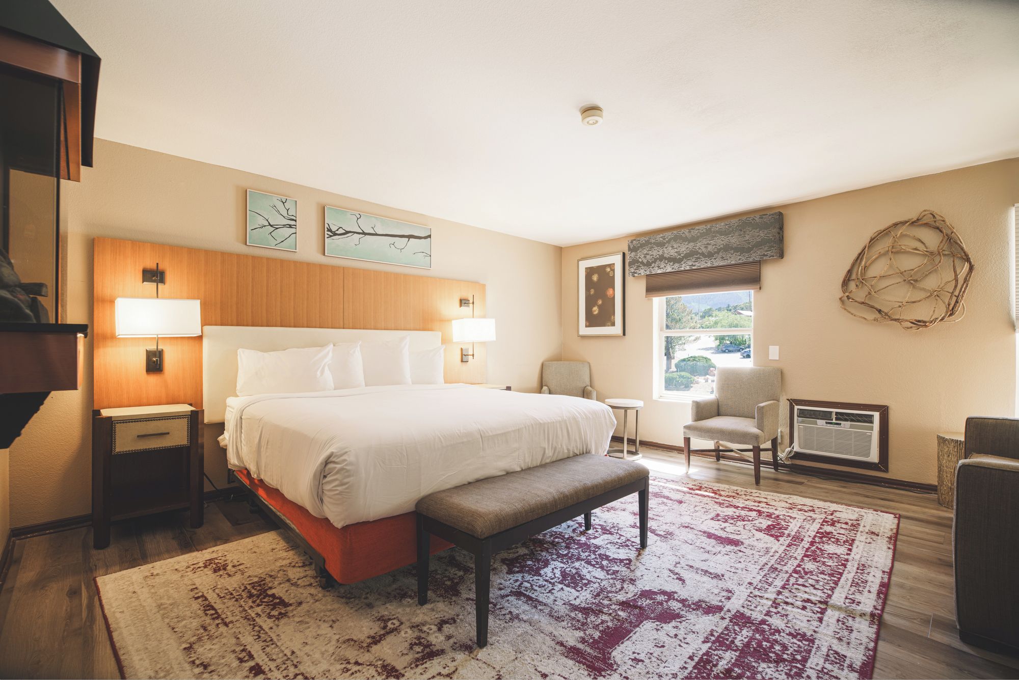 A hotel room with a large bed, bedside tables, a bench, a chair, a small table, a radiator, and artwork on the walls.