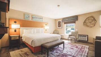 A hotel room with a large bed, bedside tables, a bench, a chair, a small table, a radiator, and artwork on the walls.