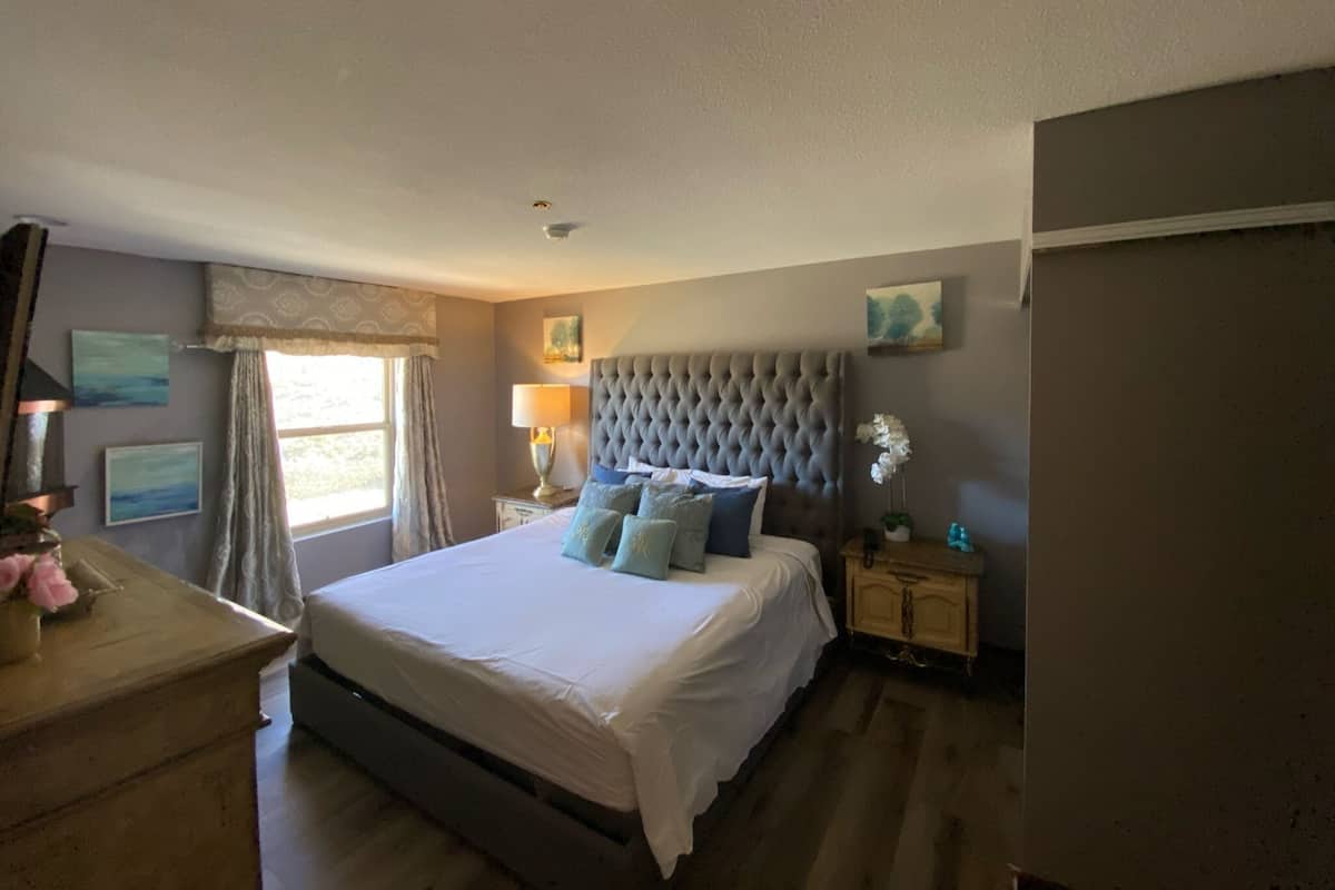 Un dormitorio con una cama grande, una mesita de noche, una lámpara y una ventana con cortinas.