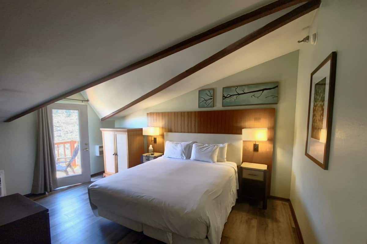 Un dormitorio con una cama grande, cabecera de madera, dos mesitas de noche con lámparas, un armario y vistas al exterior a través de una puerta.