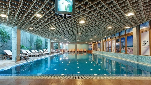 Piscina interna com água cristalina, espreguiçadeiras e uma televisão fixada no teto.