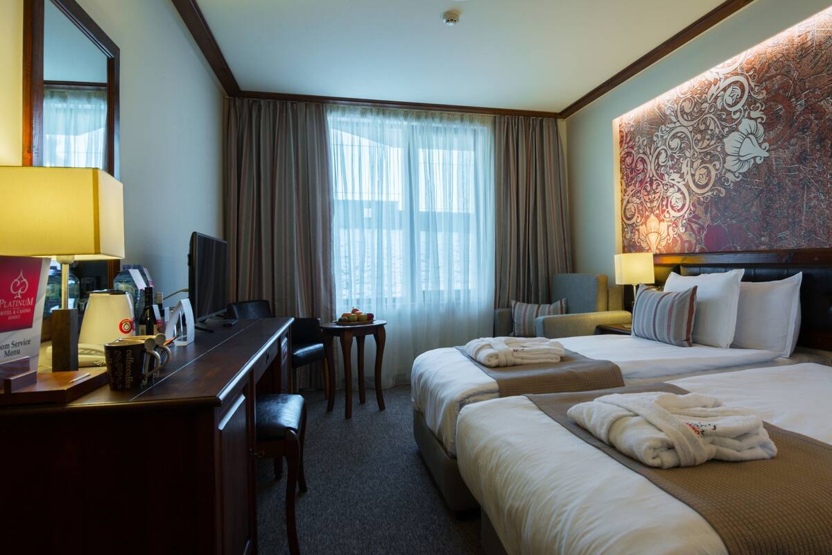 A hotel room with a desk, two beds, a large tapestry, and a view of the city.