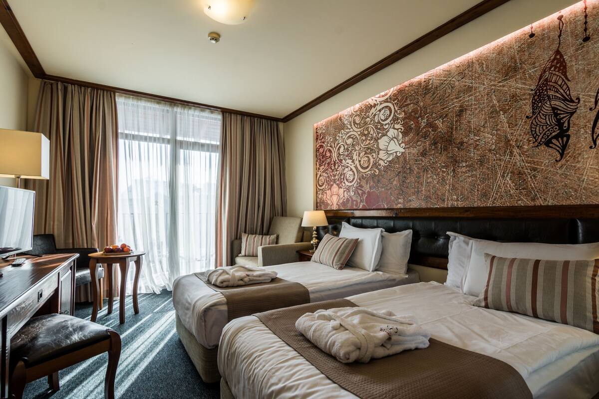 A hotel room with two beds, a desk, a chair, and a large tapestry on the wall.