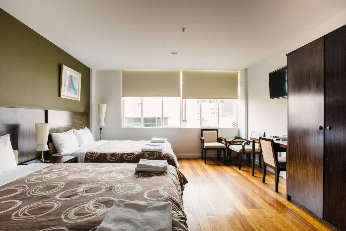 Habitación de hotel con dos camas, un escritorio, una silla, un televisor y una ventana con persianas.