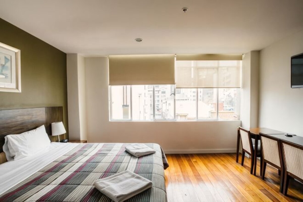 A hotel room with a large bed, a desk, a chair, a window with blinds, and a television on the wall.