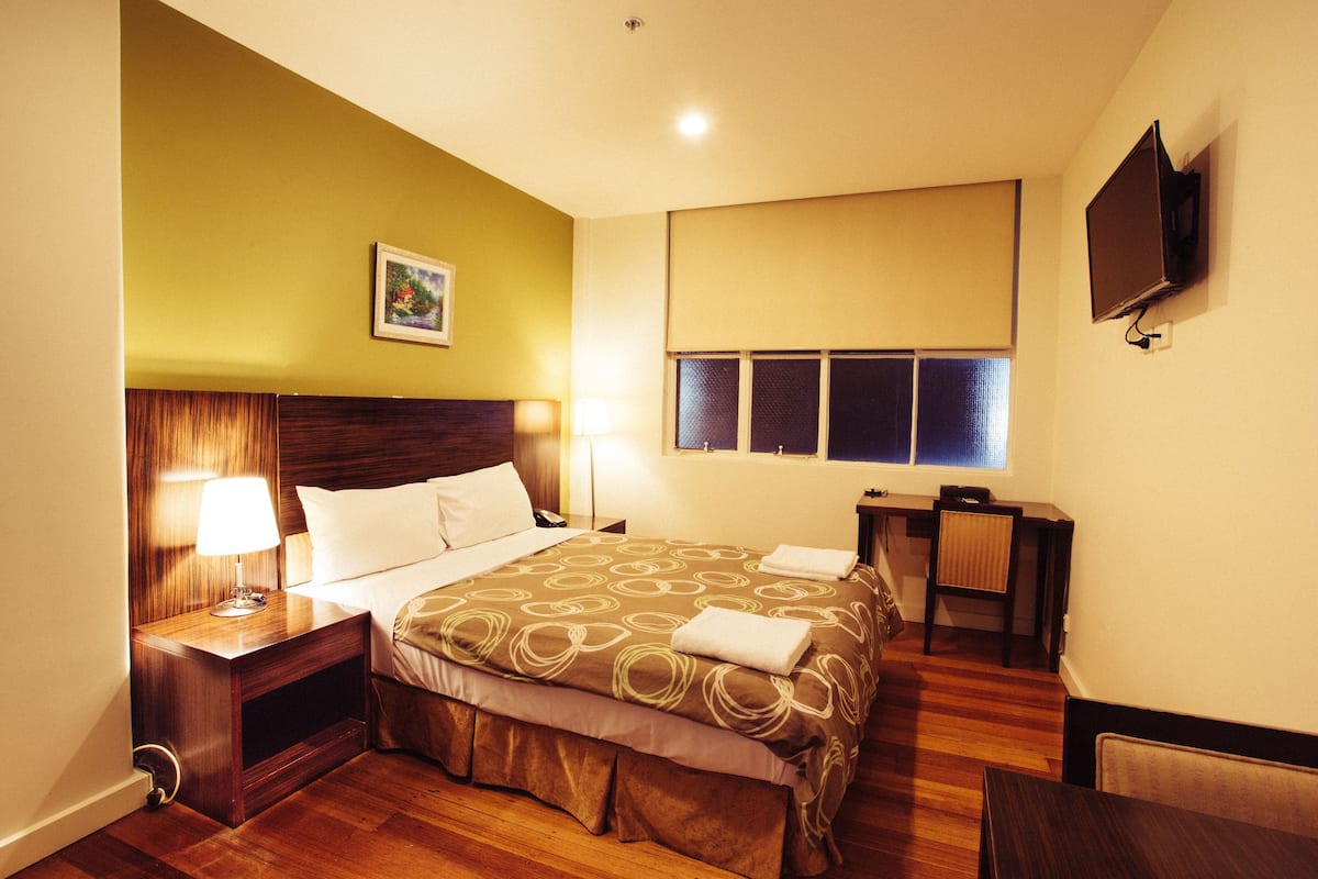 Habitación de hotel con cama, lámparas de noche, televisión, ventana con persianas y un cuadro en la pared.