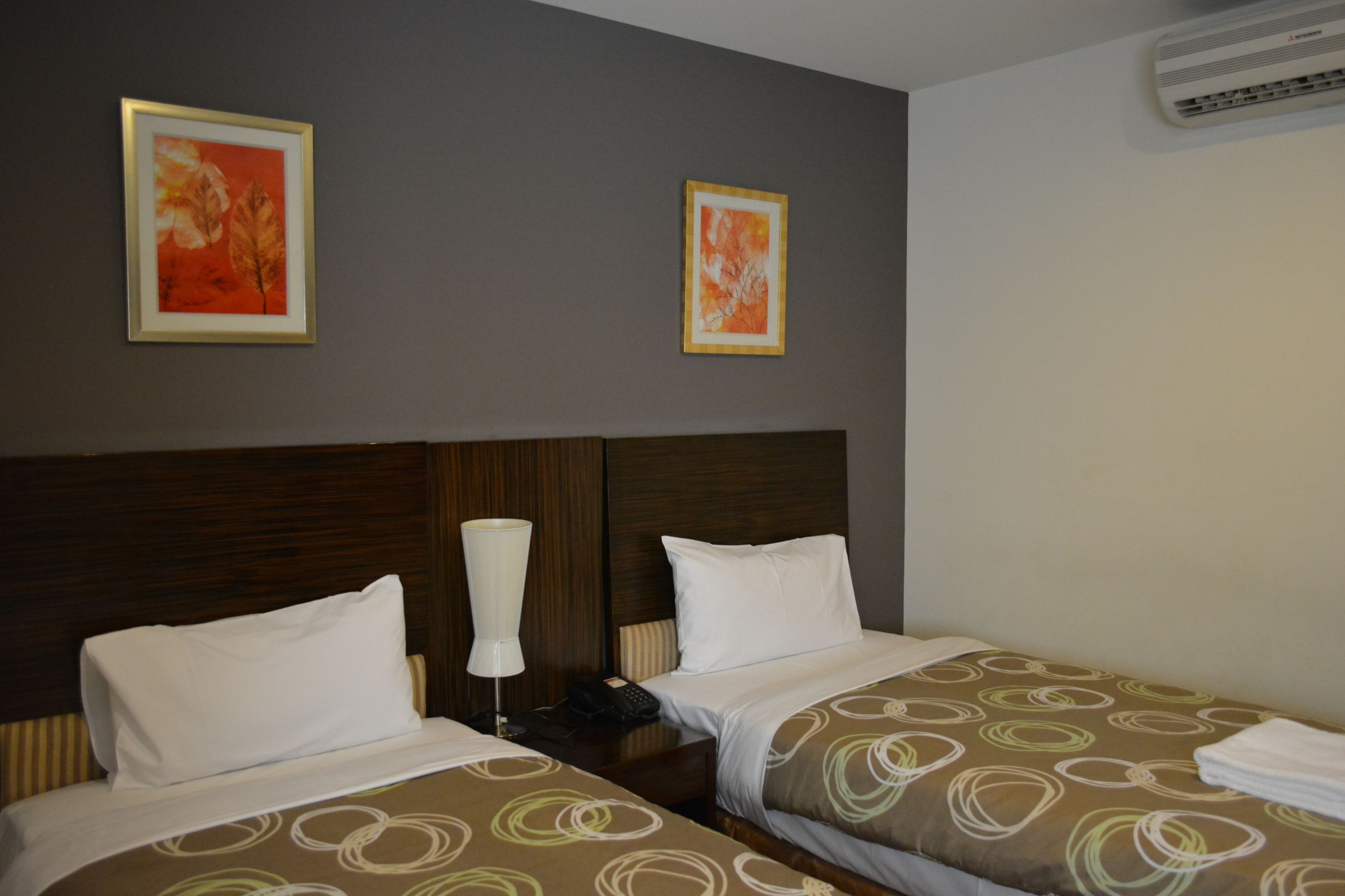 A hotel room with two beds, a wooden headboard, a bedside table with a lamp, and two framed pictures on the wall.