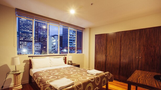 A hotel room with a large bed, a wooden wardrobe, a small table, and a window with a city view. - YTI Garden Hotel (Melbourne)