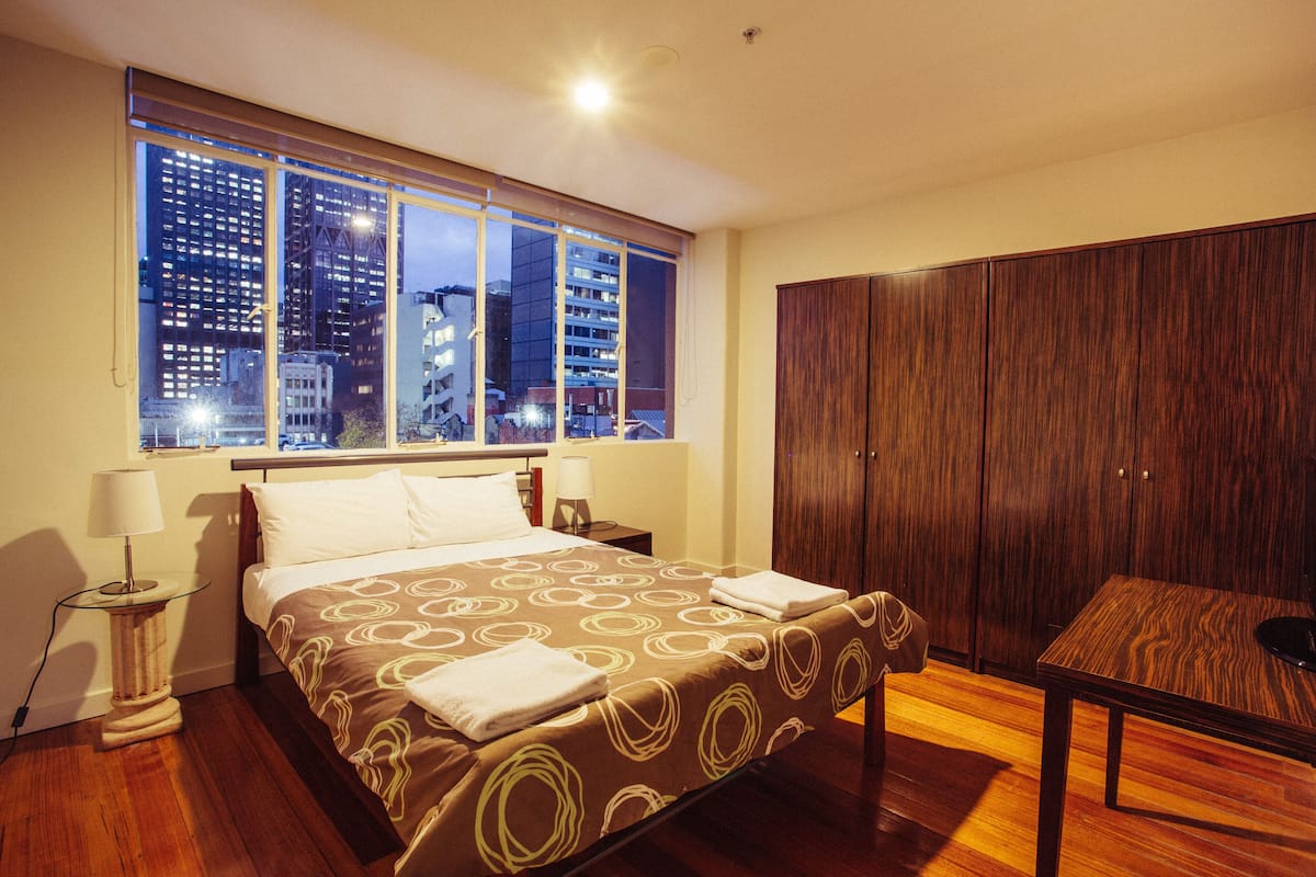 Habitación de hotel con una cama grande, un armario de madera, una mesita pequeña y una ventana con vistas a la ciudad.