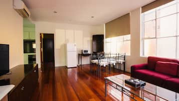 A spacious living area with a wooden floor, a glass coffee table, a red sofa, a dining table with chairs, a kitchen area with cabinets, and a TV on a cabinet.