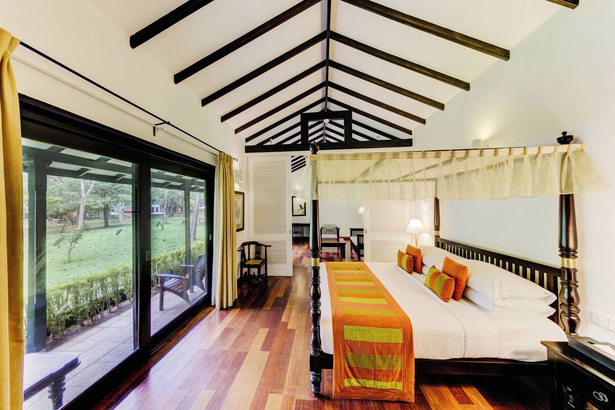 A spacious bedroom with a large bed, a wooden ceiling, and a view of a garden through a sliding door.