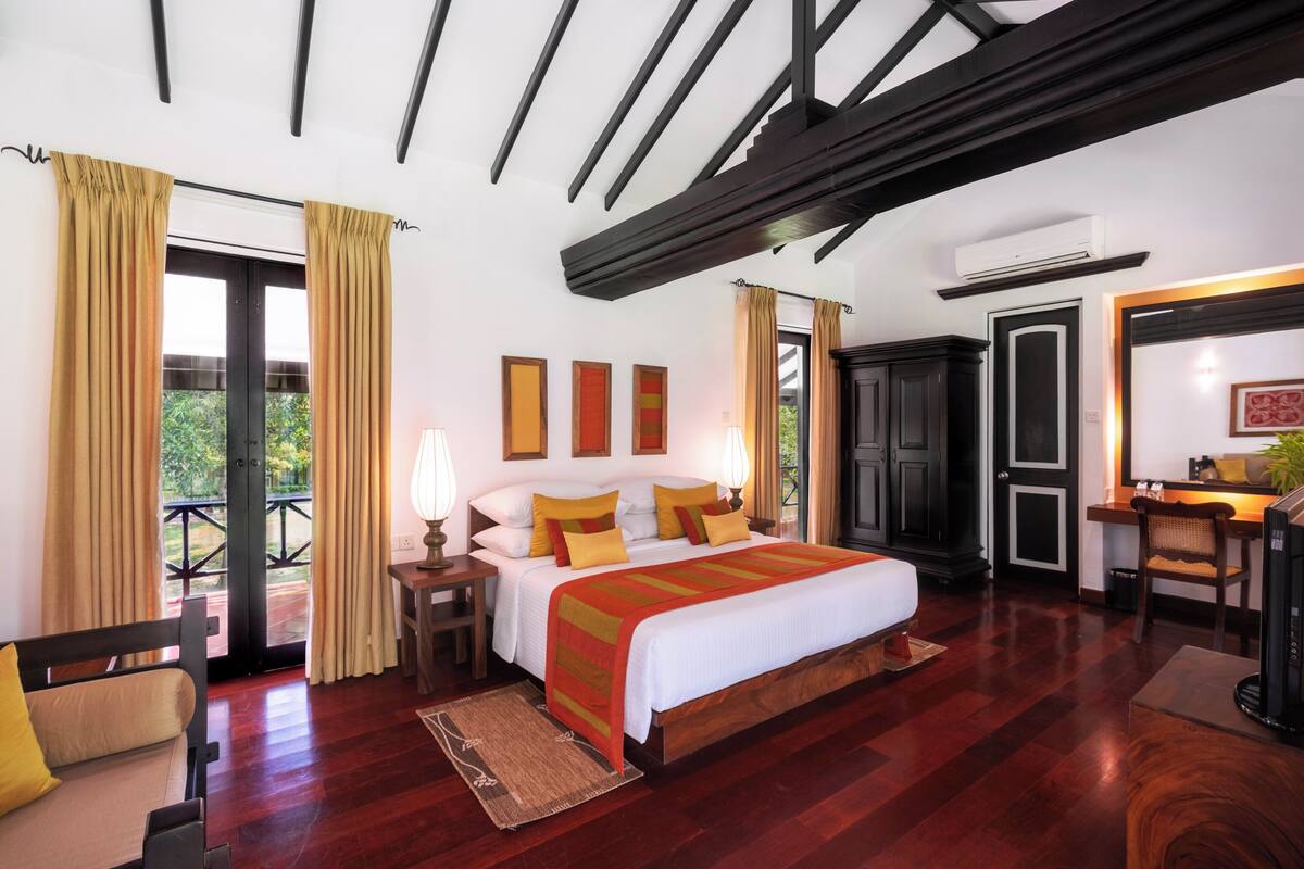 A bedroom with a large bed, wooden flooring, a desk, and a balcony with curtains.