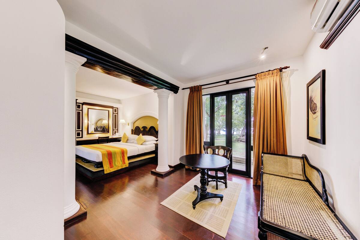 A hotel room with a bed, a dining table, a sofa, and a large window with curtains.