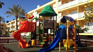 A playground with slides and climbing structures, surrounded by a wooden fence, with a yellow building in the background. - Terralta (Benidorm)