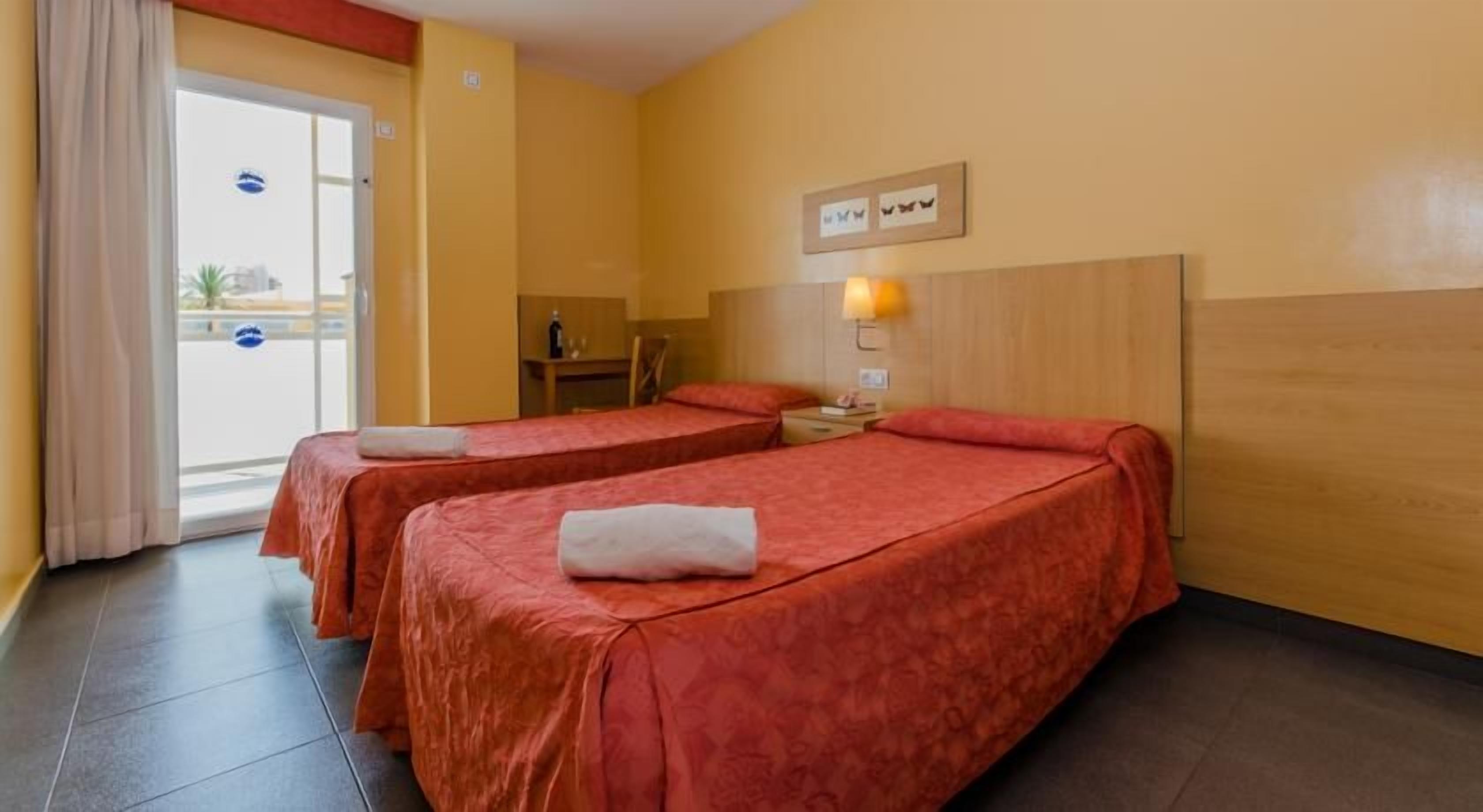 A hotel room with two beds, a wooden headboard, a nightstand with a lamp, and a view of the outside through a glass door.