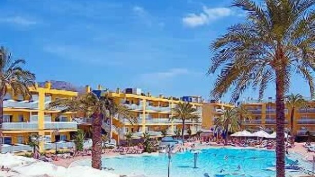 A resort with a large swimming pool surrounded by palm trees and umbrellas, multiple floors of yellow buildings, and a clear blue sky. - Terralta (Benidorm)