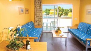A room with two blue sofas, a wooden table with a bottle and a plant, and a view of a pool and greenery outside. - Terralta (Benidorm)