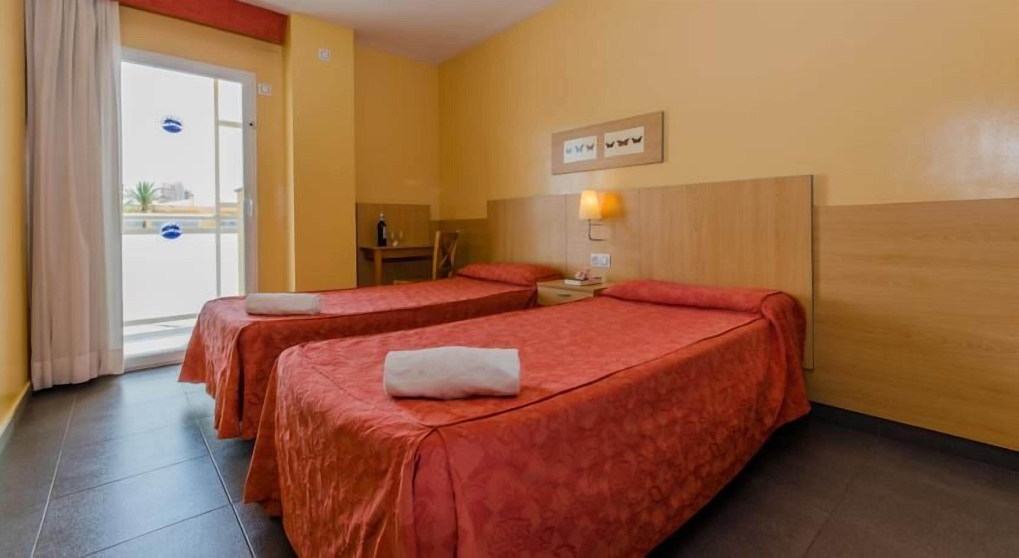 Una camera d'albergo con due letti, una testiera in legno, un comodino con una lampada e vista all'esterno tramite una porta vetrata.