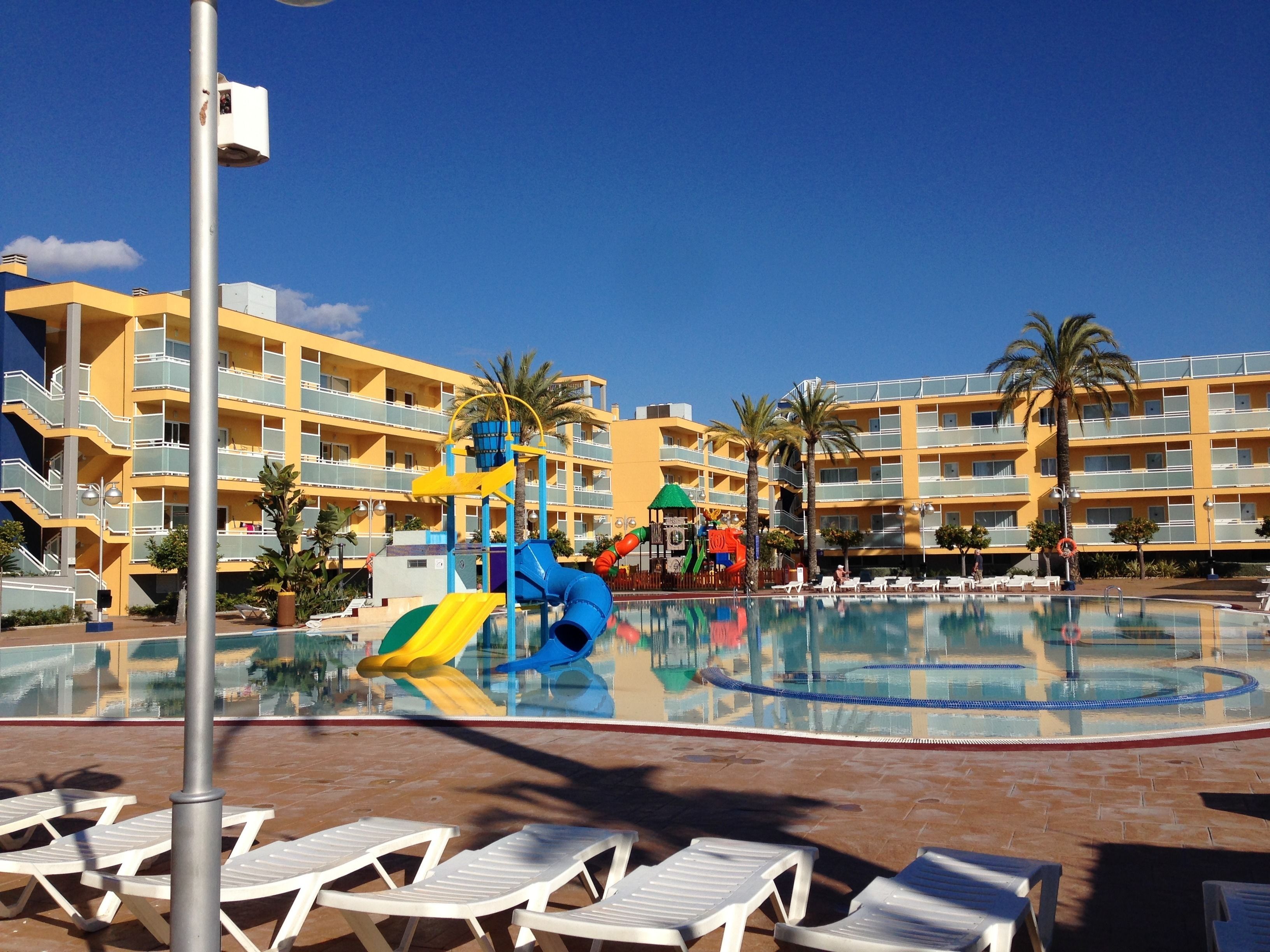 A hotel pool area with a water slide, lounge chairs, and a multi-story building in the background.