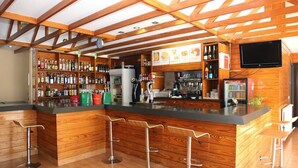 Zona de bar con vigas de madera, una barra con taburetes, estanterías con botellas y un televisor.