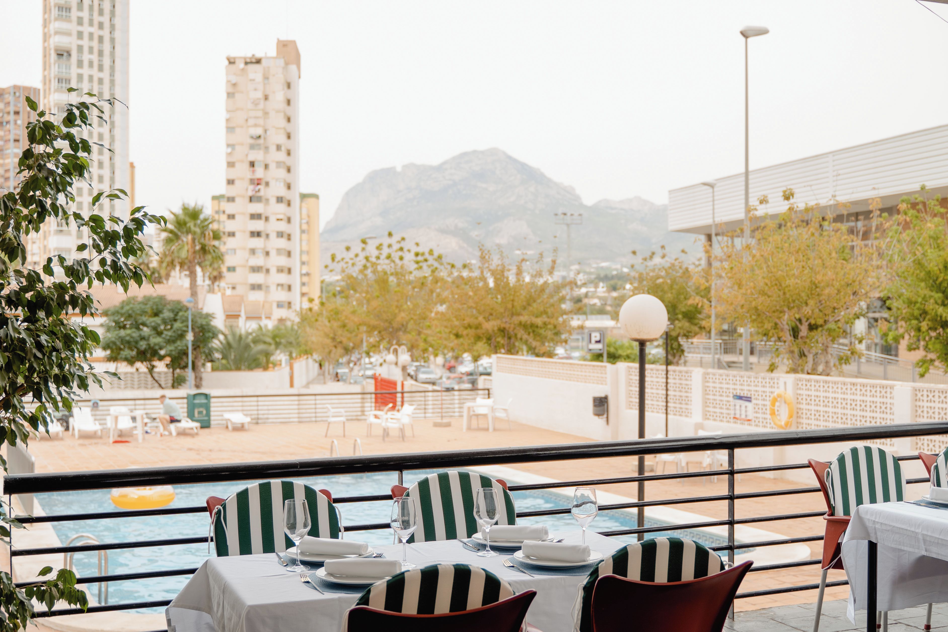 An outdoor dining area with tables set for two, overlooking a pool and a cityscape with mountains in the distance.