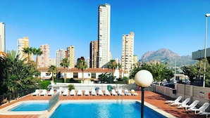 Una piscina con sillones de descanso, un paisaje urbano con edificios altos y una montaña al fondo.