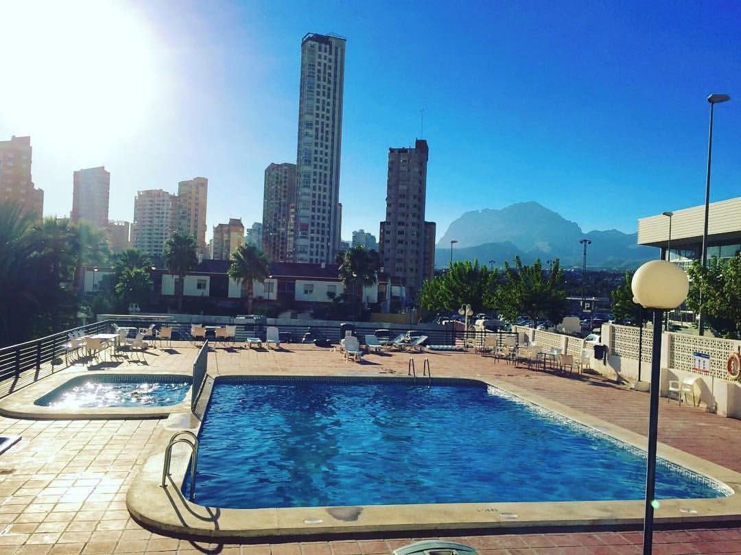 A swimming pool area with multiple pools, lounge chairs, and a cityscape with tall buildings in the background.