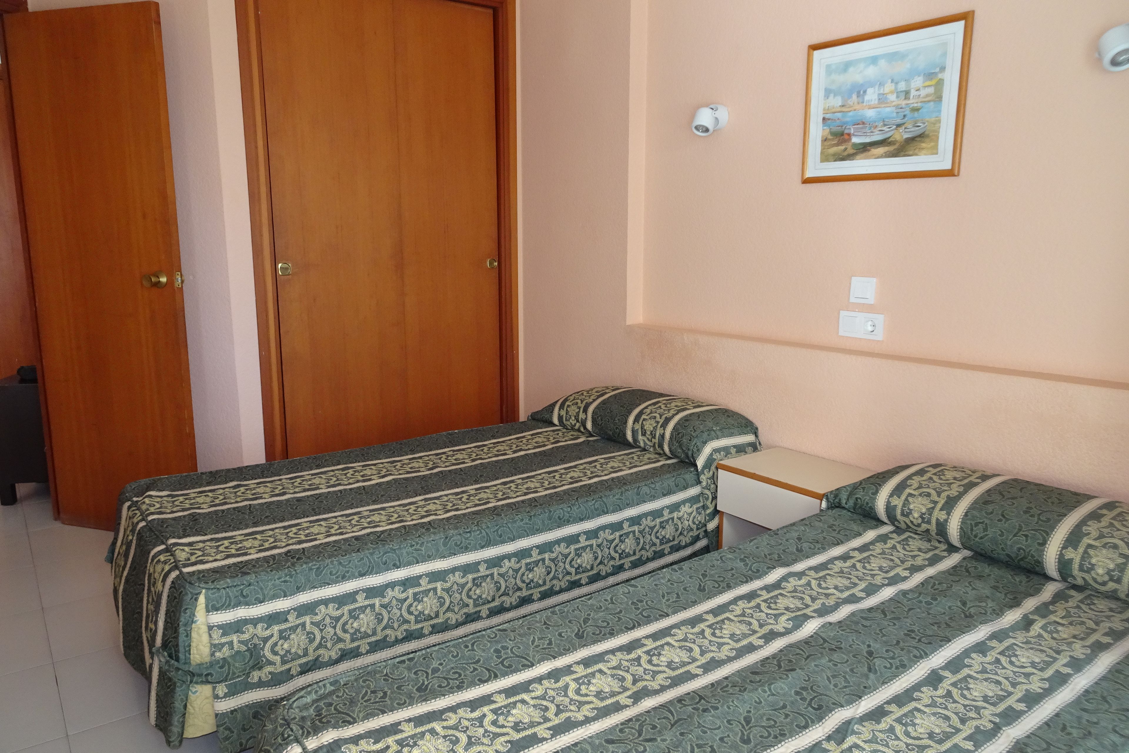 A room with two beds, a wooden door, a wall-mounted light, and a framed picture.