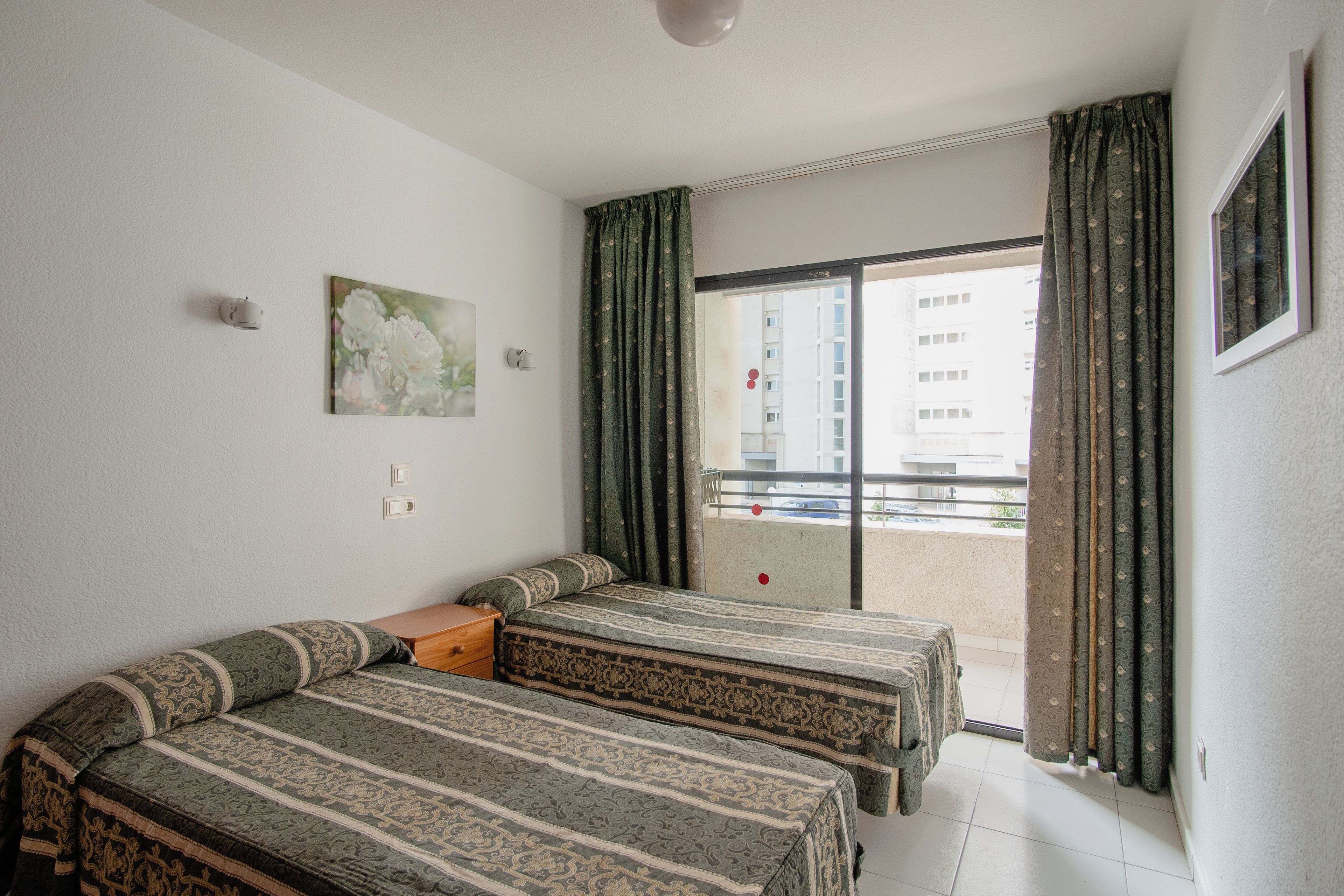 A room with two beds, a small table, a painting, and a balcony with curtains.