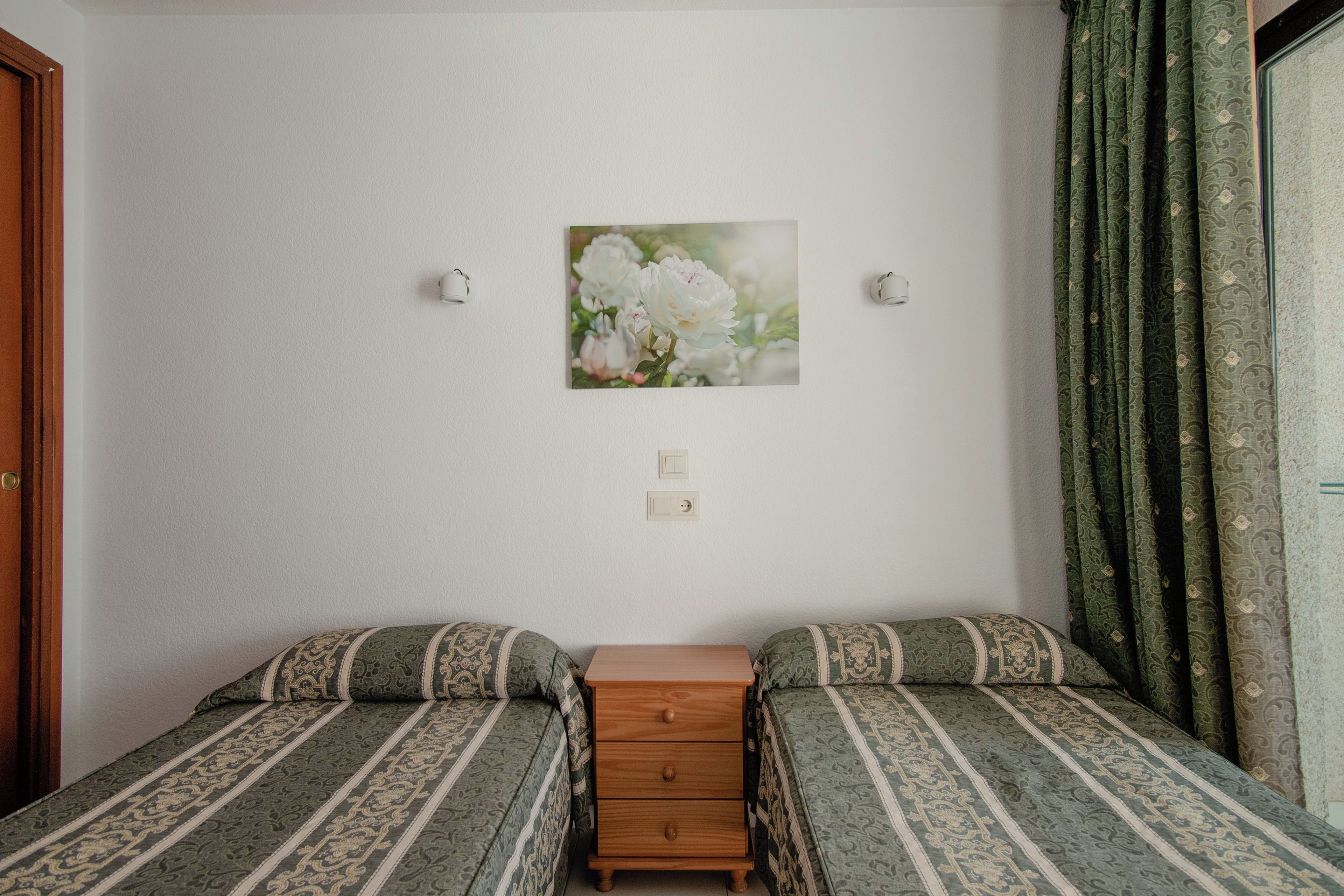 A bedroom with two single beds, a wooden nightstand, a wall-mounted light, and a floral painting.
