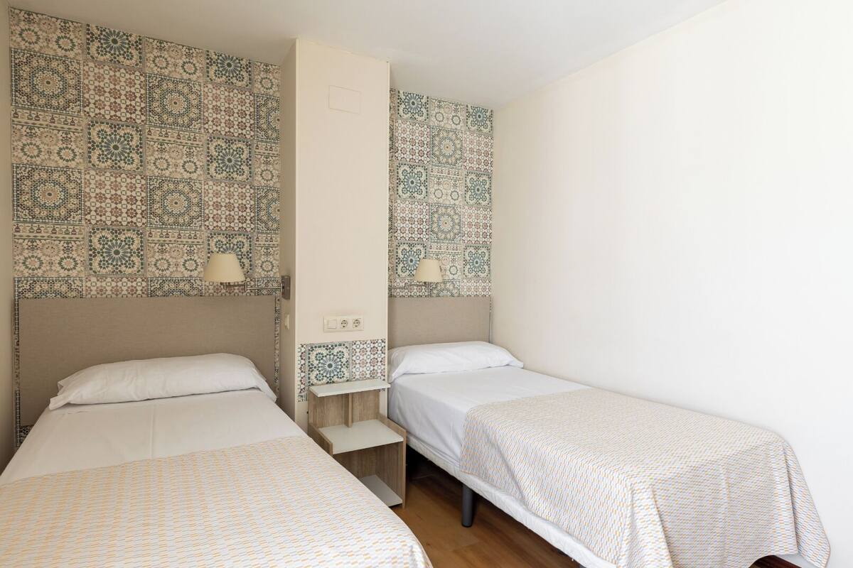 A room with two single beds, a small bedside table, a wall-mounted shelf, and patterned tile walls.