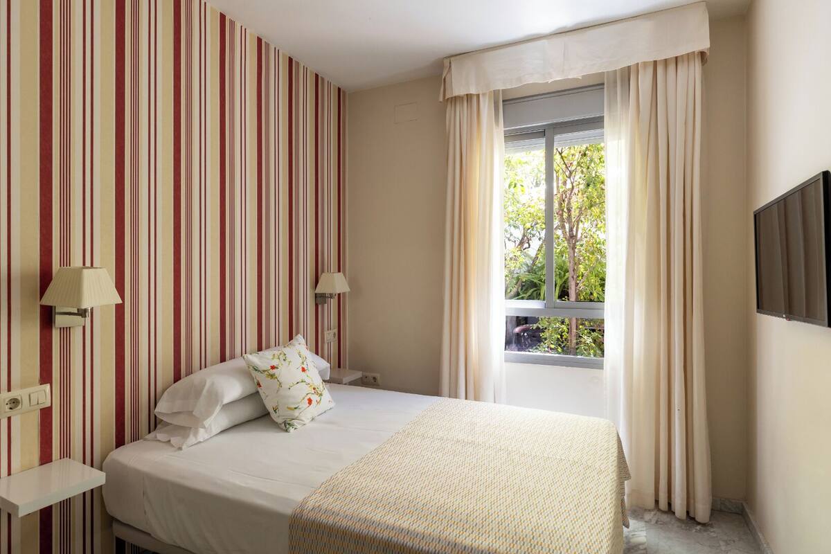 A hotel room with a bed, striped wallpaper, two bedside lamps, a window with curtains, and a TV on the wall.