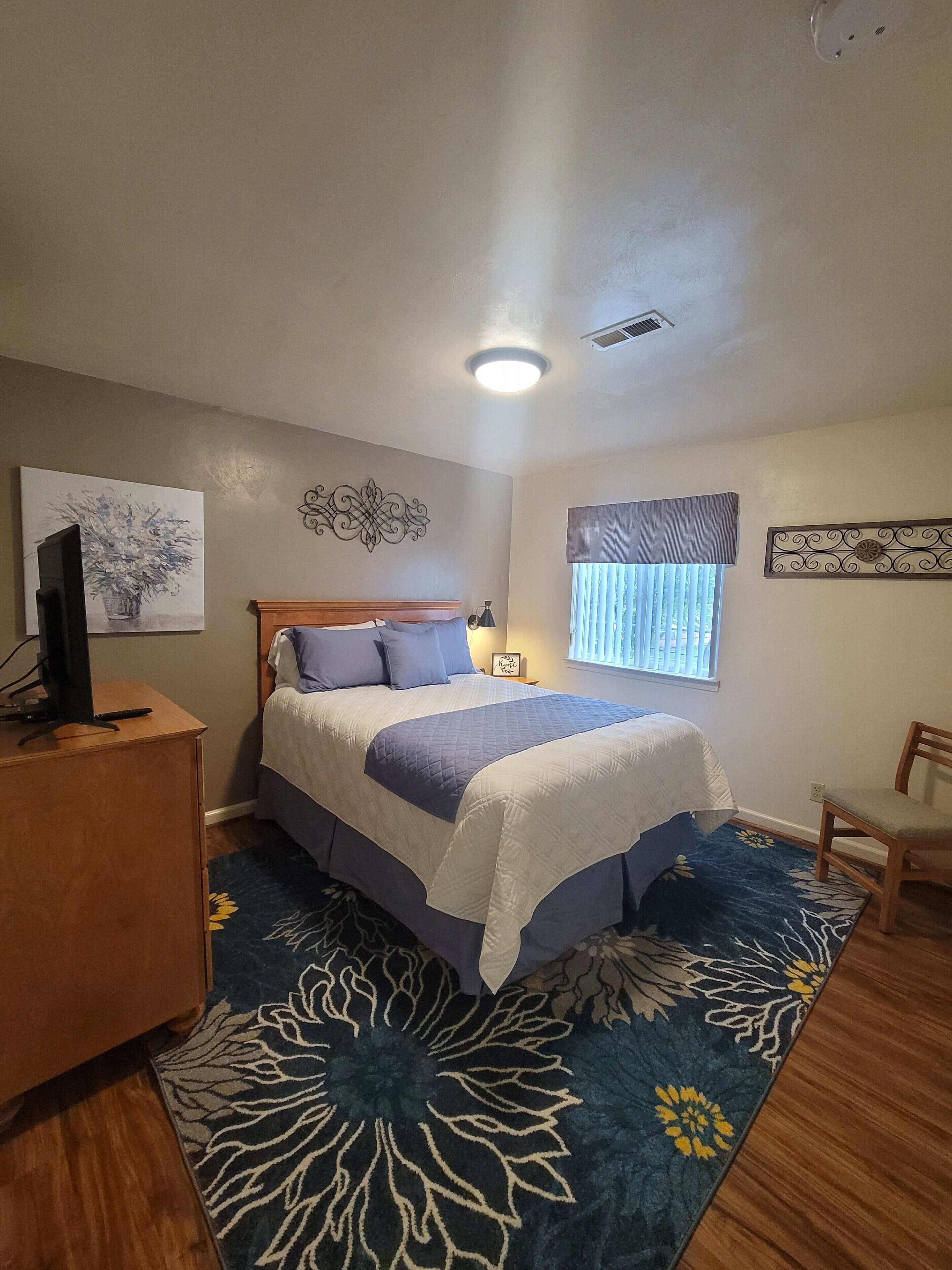 A bedroom with a bed, a desk, a chair, a television, and a window with blinds.